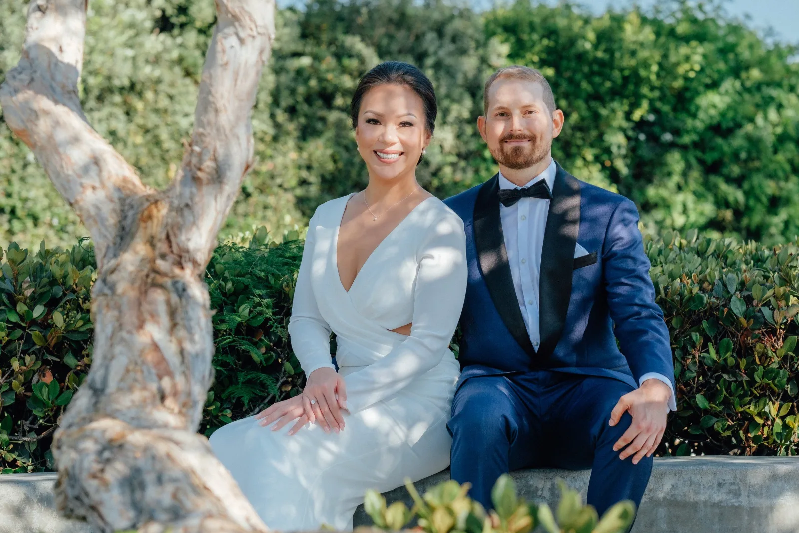 Bride in a white gown and groom in a navy tuxedo seated in a garden, Orange County wedding.