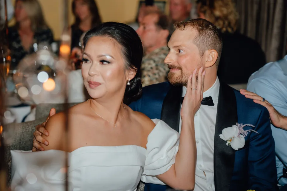 Newlyweds at a candlelit reception, bride in an off-shoulder gown and groom in a navy tuxedo, Orange County wedding.