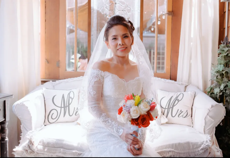 Bride in a lace off-shoulder gown seated with a bouquet in a sunlit getting-ready room, Orange County wedding.
