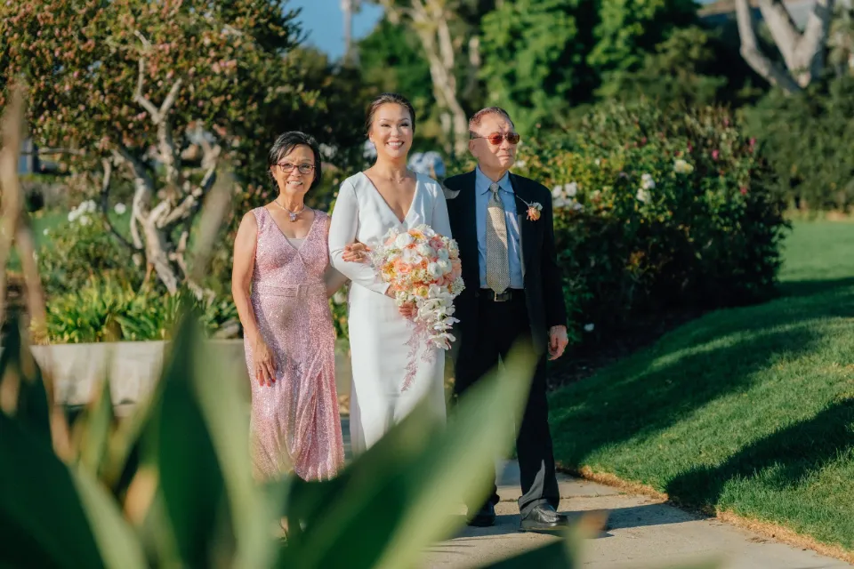 Bride walking a garden path with her parents, holding a peach and white bouquet, Orange County wedding.