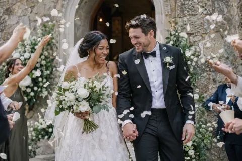 Newlyweds laughing as guests toss petals outside a stone chapel